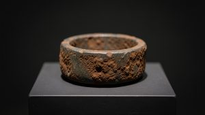 Bague funéraire grecque antique en fer corrodé présentée sur un socle de musée sombre sous un éclairage scientifique doux.